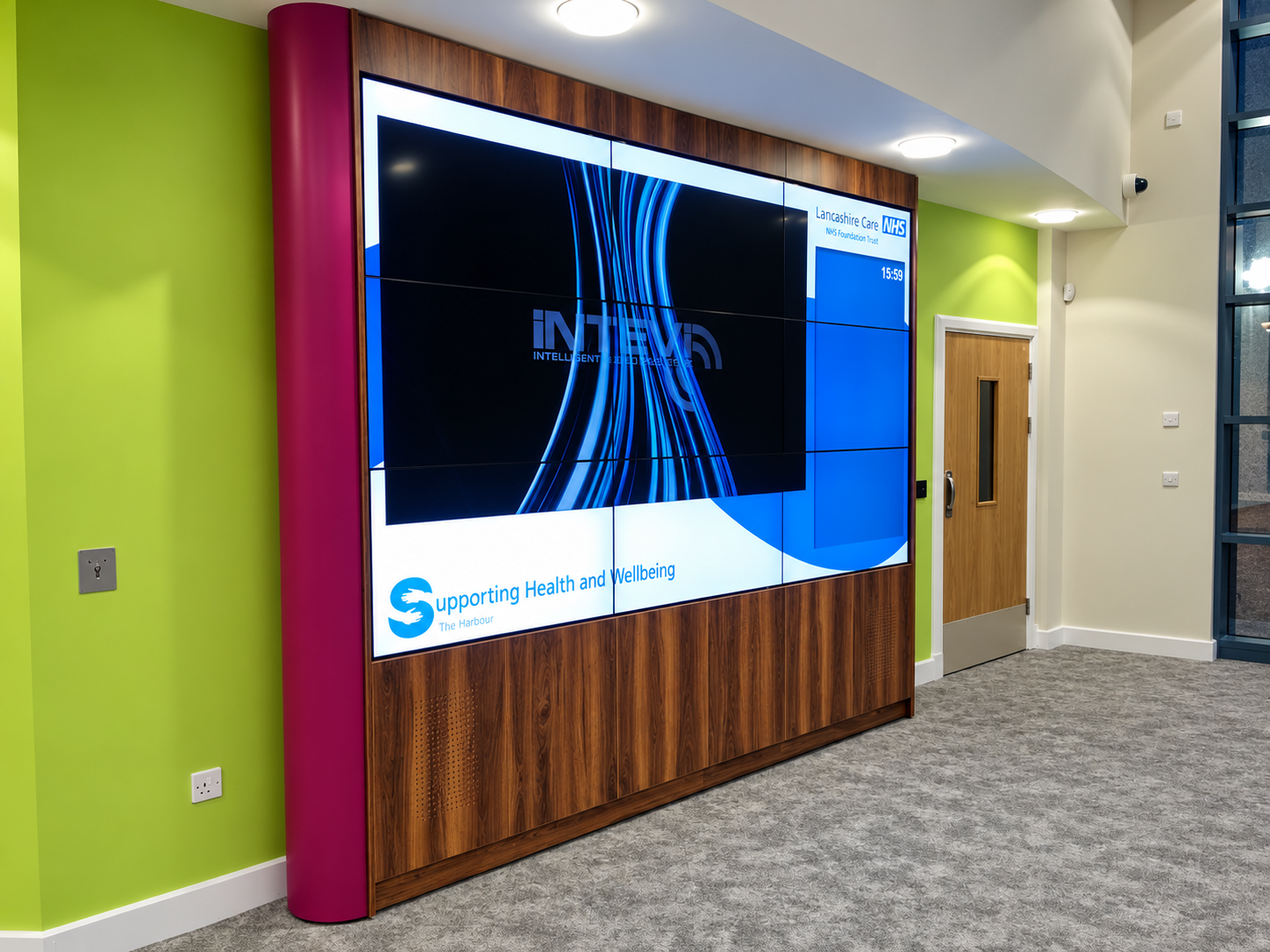 Large NHS video wall set within a wood-grain surround with a curved magenta side panel beside a lime green wall.