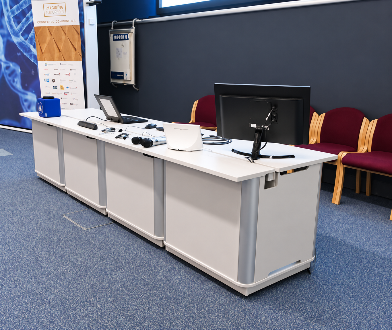 Long light grey AV teaching desk with a white worktop, monitor, laptop and microphones, with burgundy chairs behind.