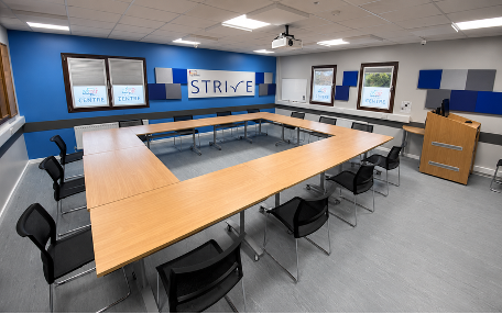 U-shaped wood-finish training tables with black chairs, a matching lectern and blue wall panels in a STRIVE training room.