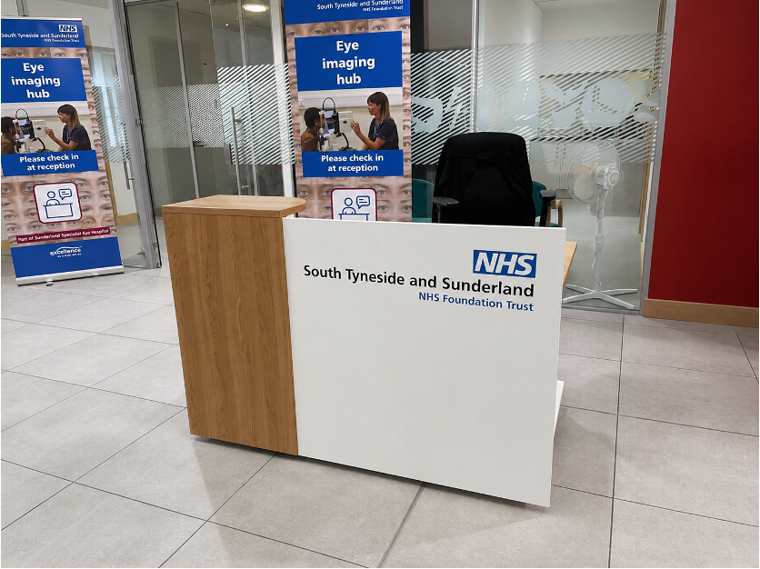 White and oak-effect reception desk with South Tyneside and Sunderland NHS Foundation Trust branding at an eye imaging hub.